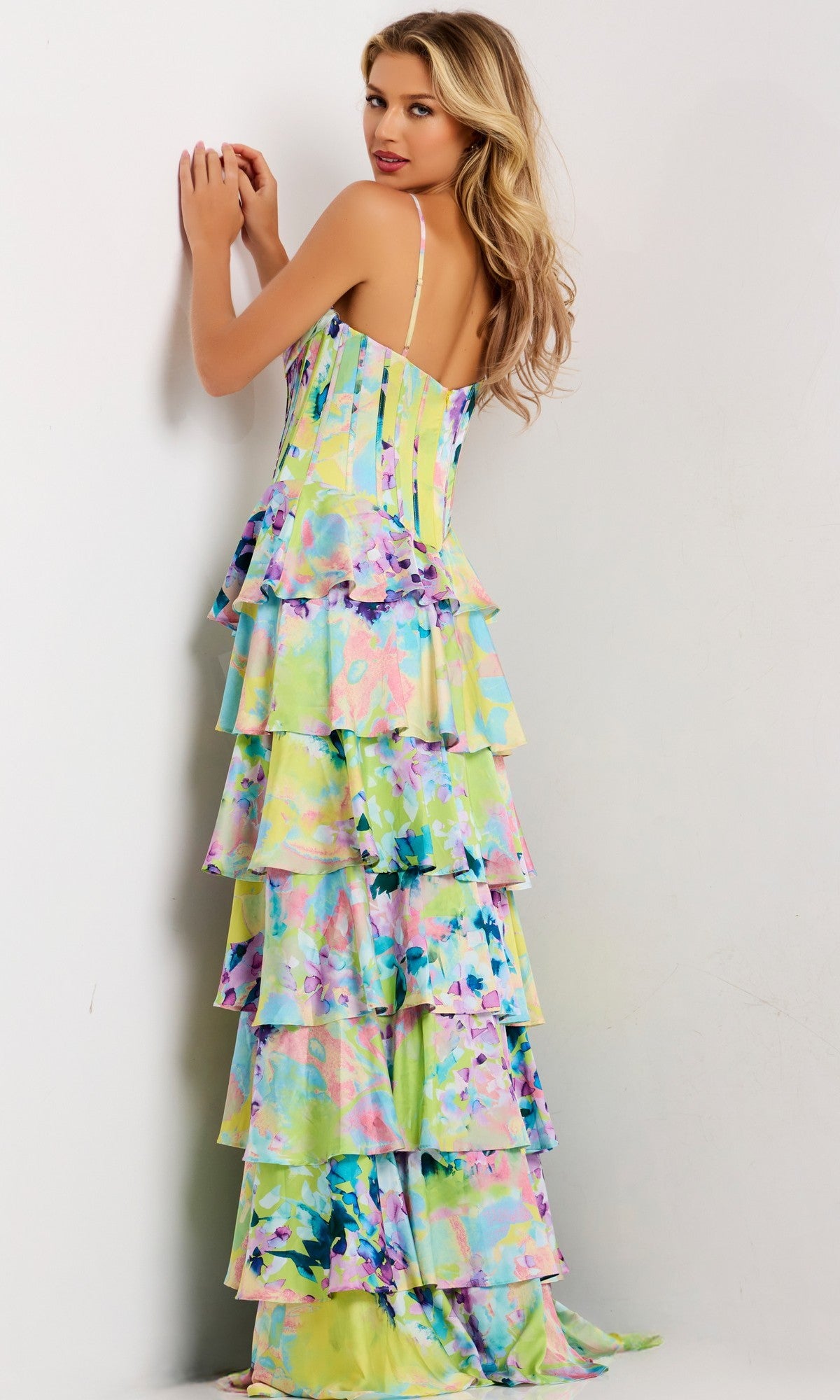 A woman stands against a white wall wearing the Jovani 47560 Long Prom Dress, featuring thin straps and pastel, multi-tiered ruffles—an elegant look with a watercolor-inspired design.
